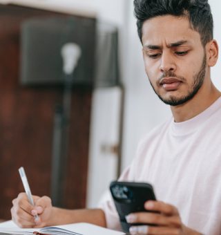 Um estudante olhando para o seu celular em uma mão. Ele está sentado e com a outra mão escreve em seu caderno