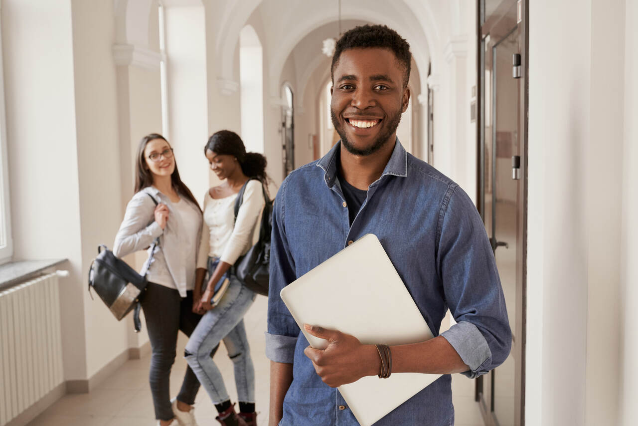 jovem estudante posa sorridente no corredor da universidade com colega ao fundo