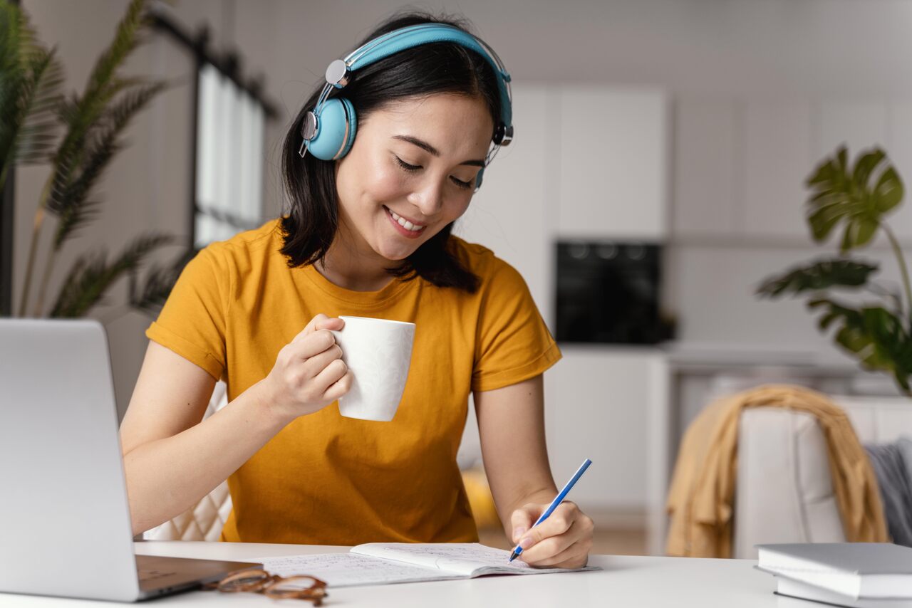 jovem faz anotações diante do computador enquanto toma café com fones de ouvido