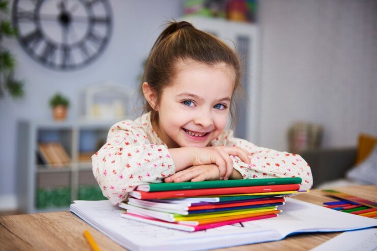 Uma pequena aluno sorri enquanto está com os braços apoiados em alguns cadernos em cima de uma mesa