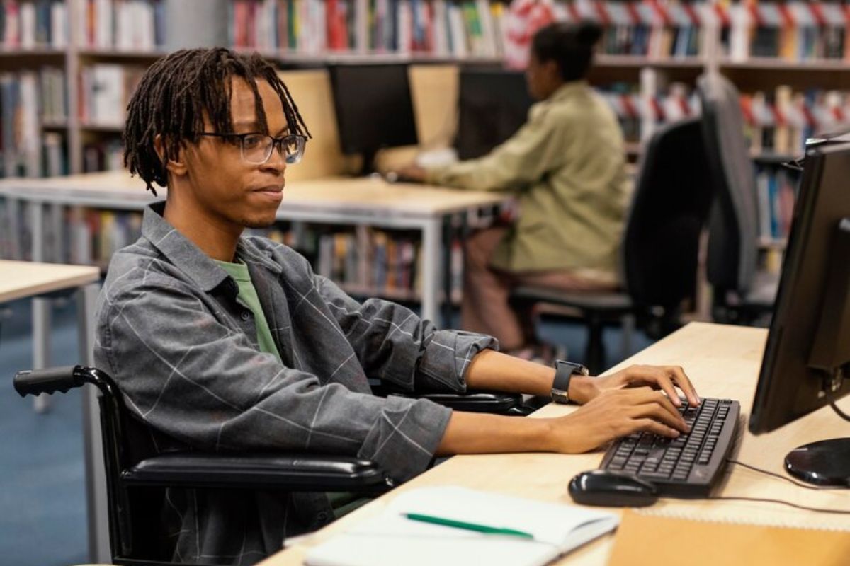 Um aluno cadeirante está dentro de uma biblioteca sentado em frente a um computador enquanto digita