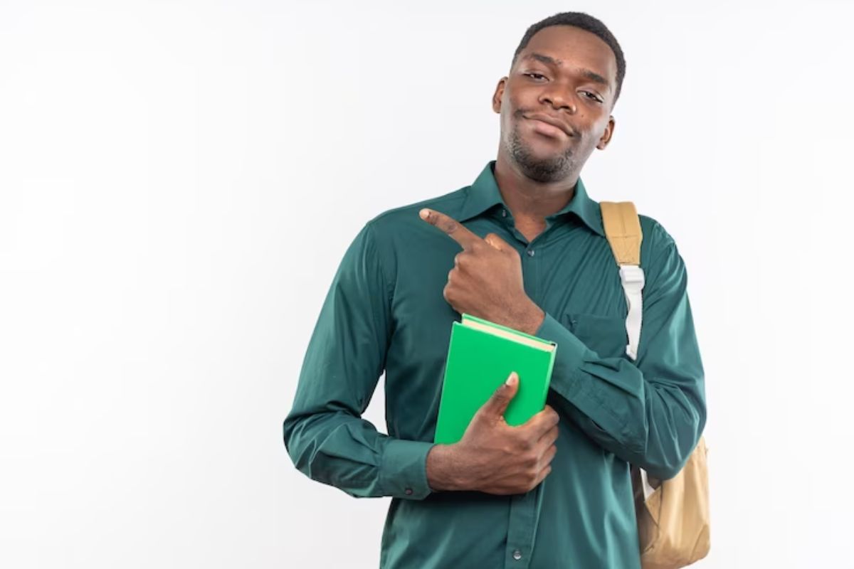 Um aluno segura um caderno verde no colo enquanto aponta para o lado segurando a mochila por uma alça