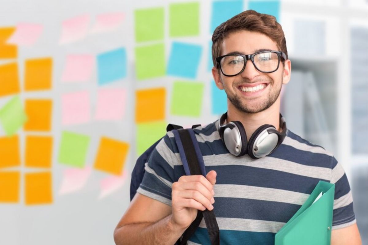 Um estudante sorrindo após aprovação no Vestibular Quero Bolsa 2024. Ele usa óculos, está com headphone no pescoço e segura uma pasta entre o braço