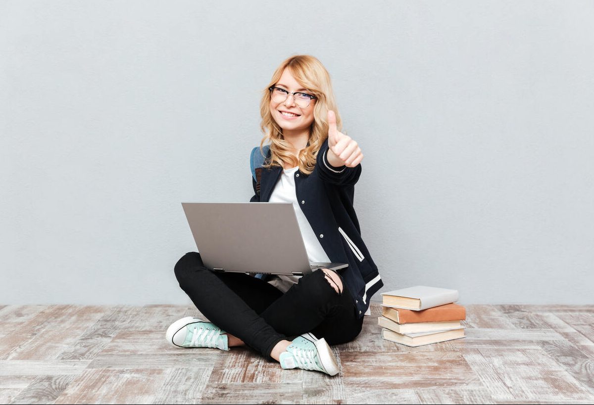 Jovem sentada no chão sorri e faz sinal de positivo com notebook em seu colo