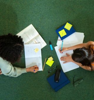 [Vista de cima] duas alunas de bruços no chão estudam e fazem anotações no caderno acerca de Como Funciona o FIES 2025