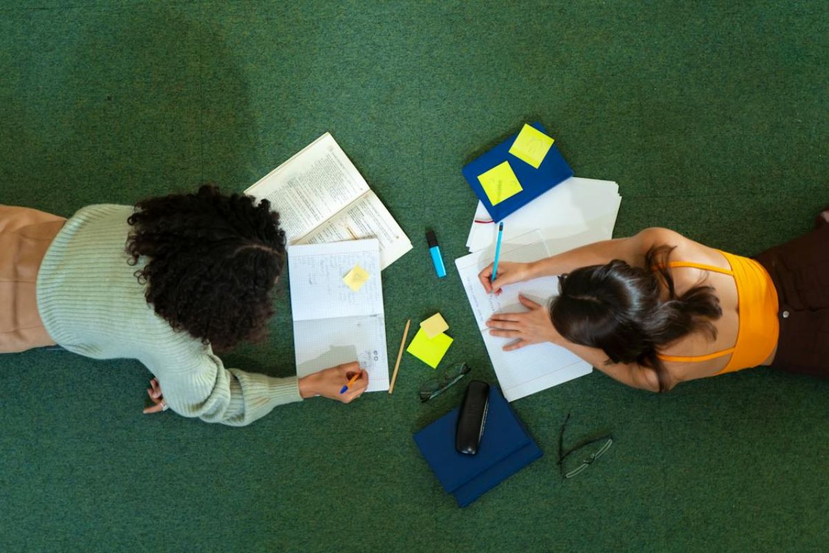 [Vista de cima] duas alunas de bruços no chão estudam e fazem anotações no caderno acerca de Como Funciona o FIES 2025