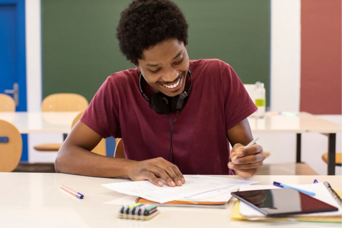 Um jovem na sala de aula sentado estudado. Ele sorri enquanto faz anotações em seu caderno