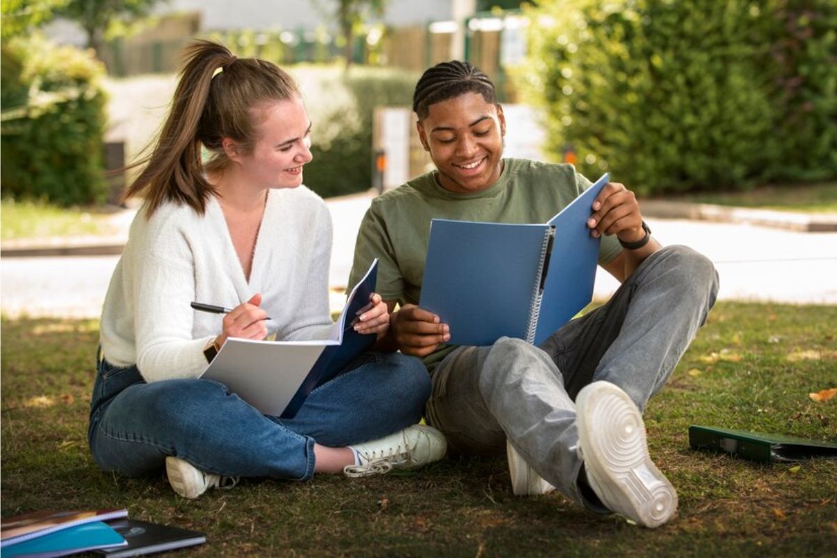 Dois alunos com seus cadernos nas mãos, conversam enquanto estão sentados na área externa da faculdade