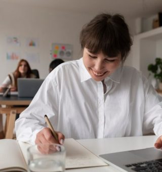 Uma estudante na sala de aula mexe em seu notebook. Ao mesmo tempo ela anota alguns dados sobre o Edital SISU 2025 em seu caderno