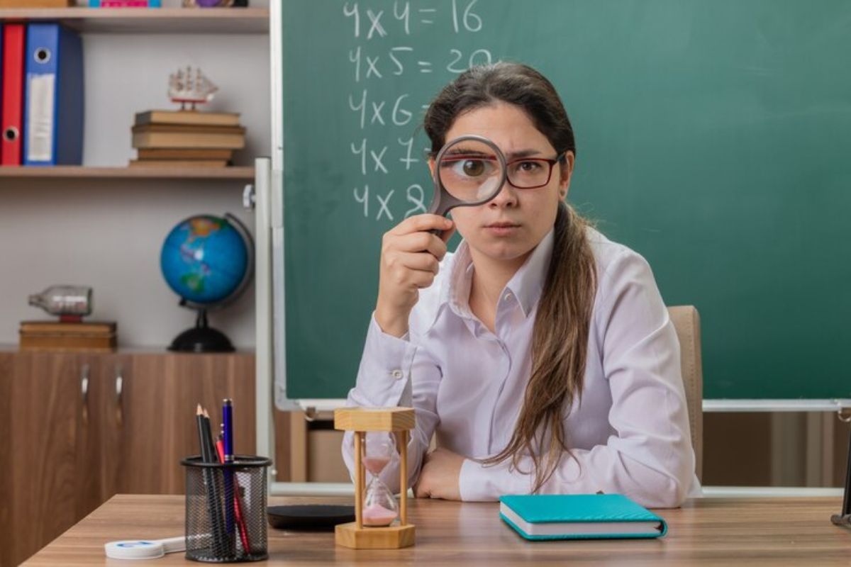 Uma professora sentada na sala de aula com uma lupa em frente ao olho