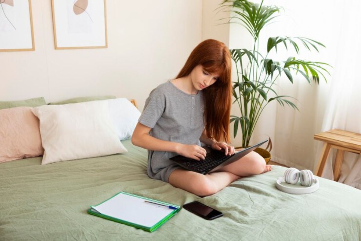 Uma menina está sentada na cama estudando com o notebook em cima das suas pernas. Ao lado, na cama, há um caderno e um celular