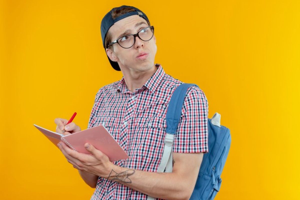Um aluno com o caderno na mão olhando para cima com olhar pensativo. Ele está fazendo anotações, usa boné e segura a mochila azul por uma alça