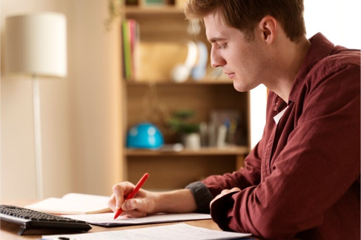 Um aluno visto de lado. Ele está anotando algo em seu caderno enquanto estuda pelo notebook