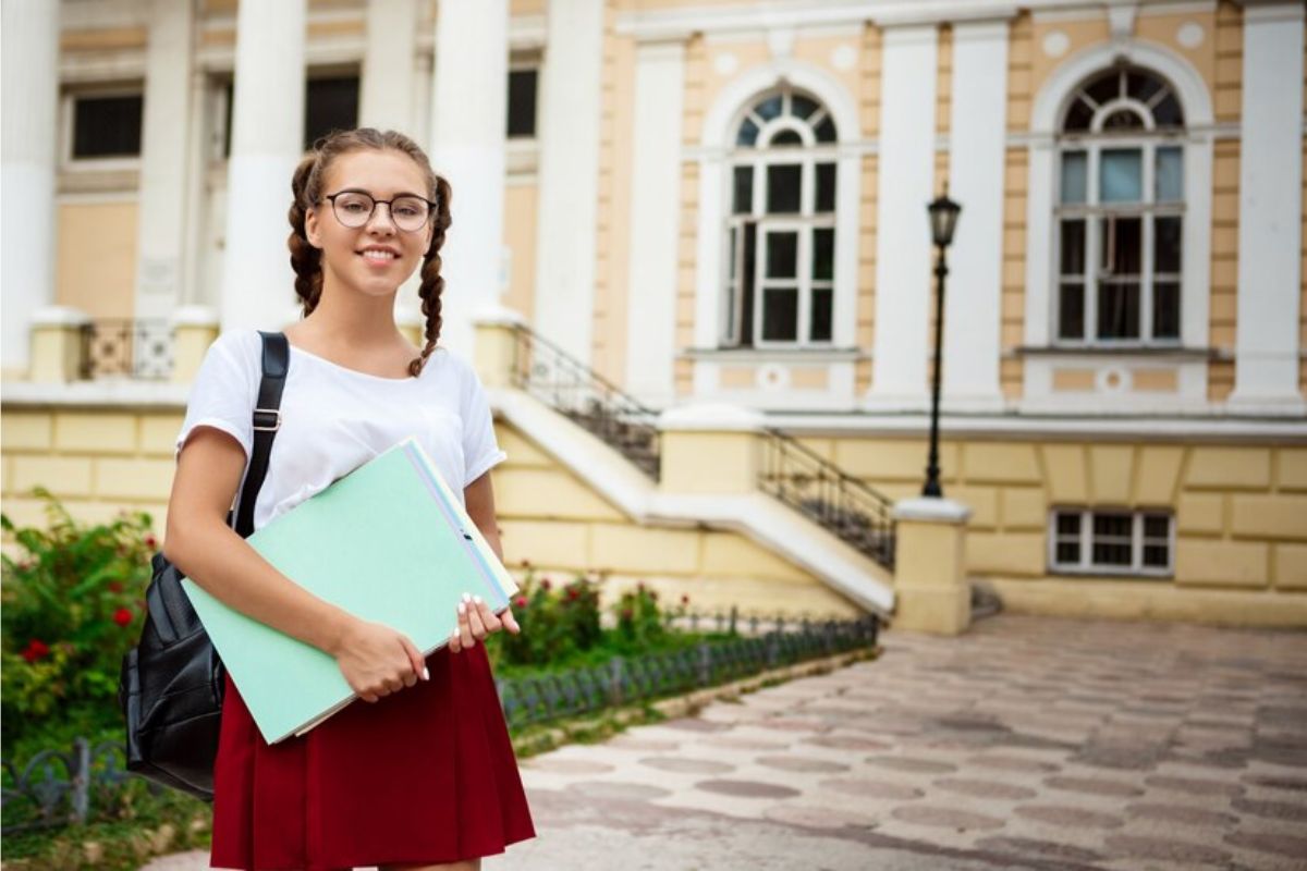 Uma aluna parada na frente da faculdade segurando seu material