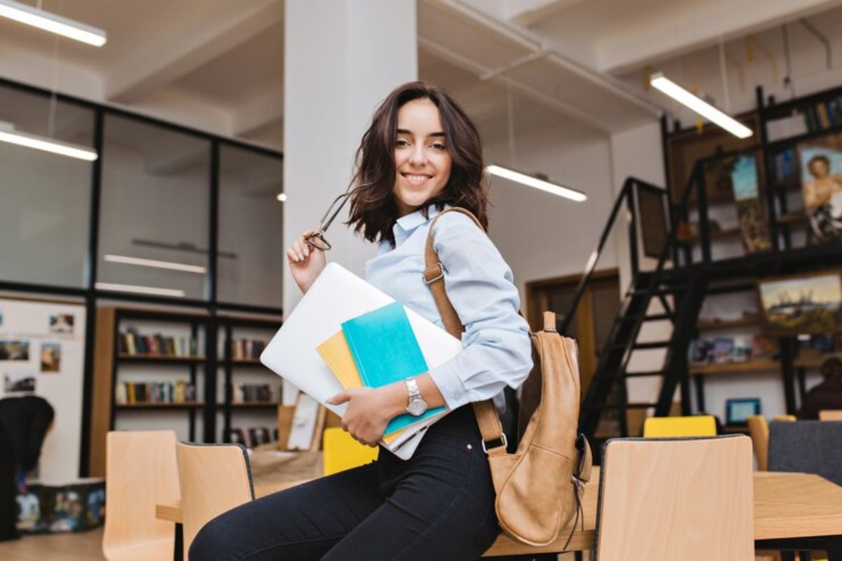 Uma aluno na biblioteca da universidade segura alguns cadernos e pastas no braço. Além disso, ela está com sua mochila e sorri para a foto