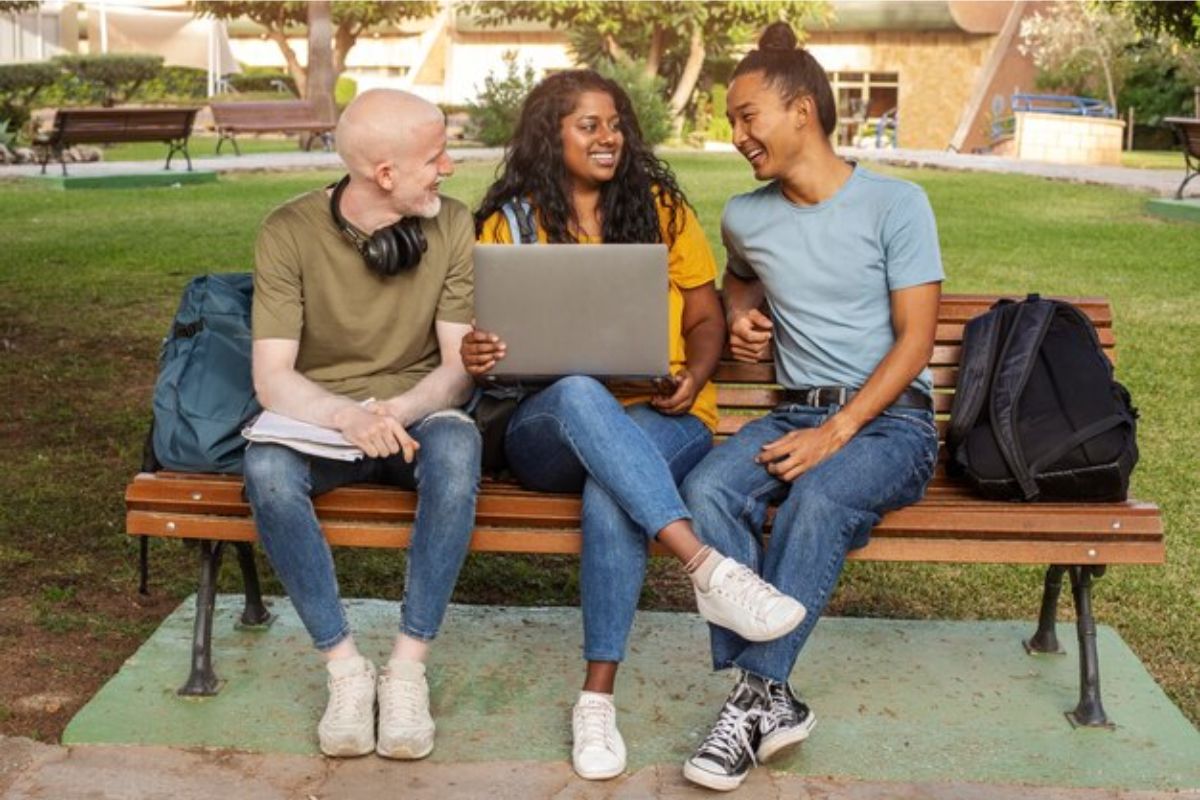 No banco da praça da faculdade. Uma aluna segura um notebook no colo, ela está no meio de outros dois amigos que conferem os Cursos do ProUni 2025
