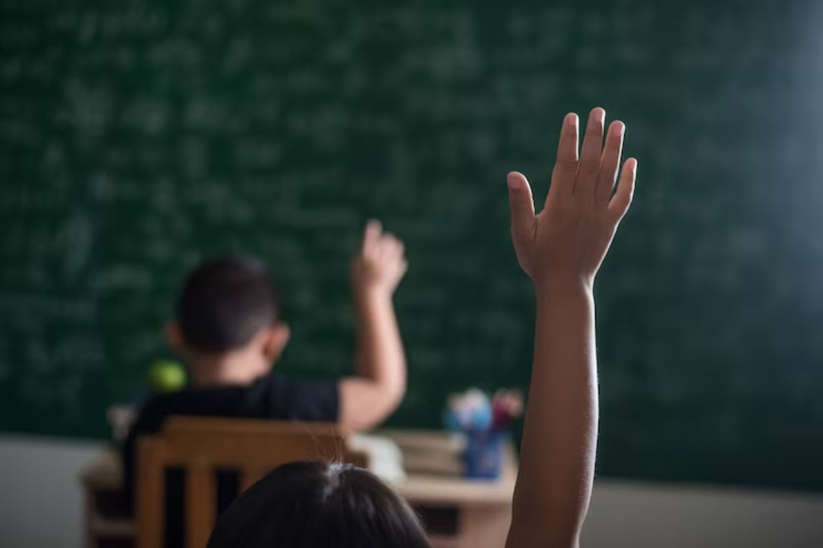 Na imagem uma criança dentro da sala de aula levanta a mão esticando o braço para fazer uma pergunta