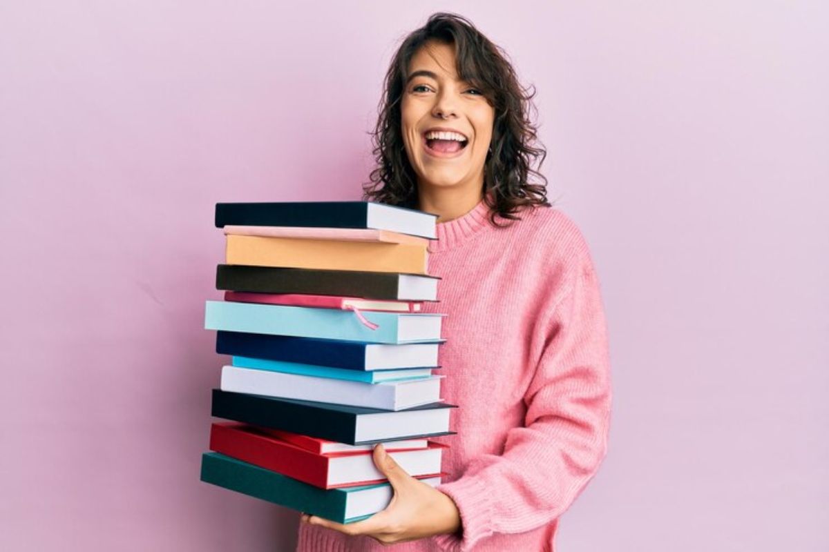 Uma mulher com um moletom rosa segura uma pilha de livros enquanto sorri