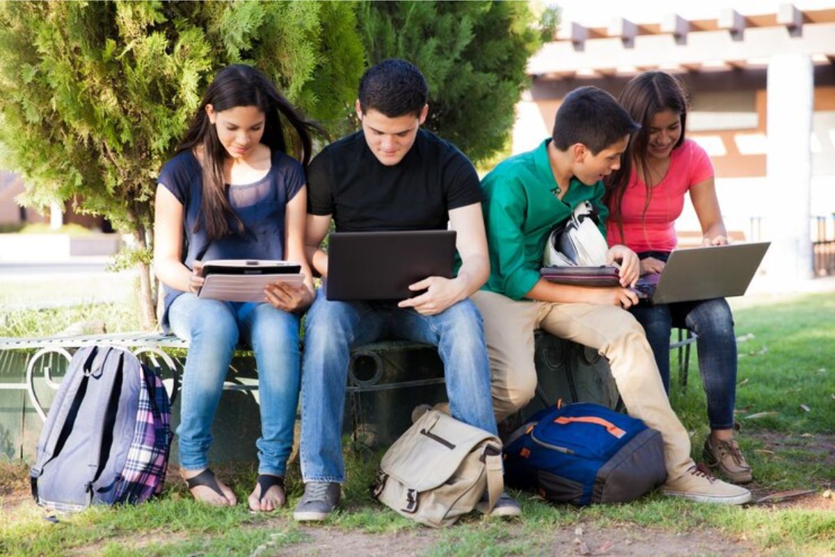 Quatro alunos estão sentados no jardim da escola. Dois alunos conferem o notebook, e outros dois pelo tablet