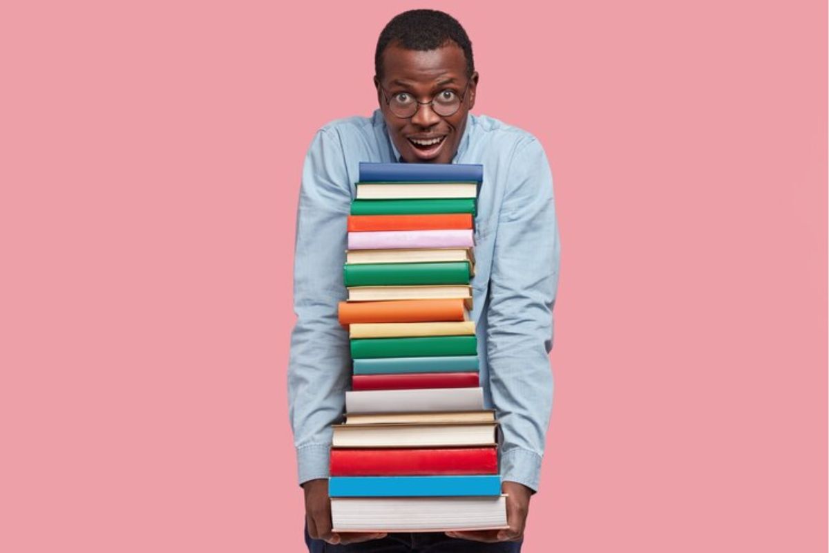 Um professor segurando uma grande pilha de livros com as duas mãos e apoiando o seu queixo. Estes são os Materiais de Apoio ao Currículo Paulista 2025