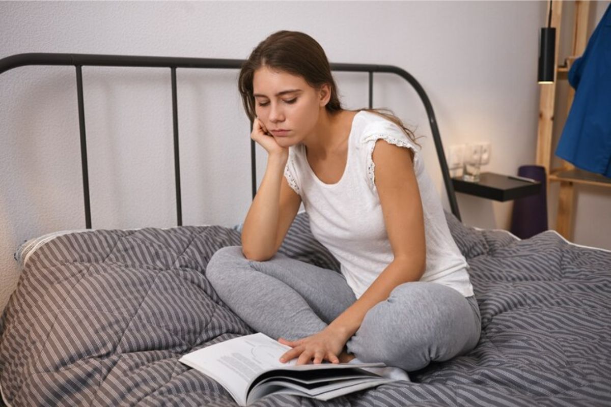 Uma menina sentada em sua cama com as pernas cruzadas enquanto lê um livro
