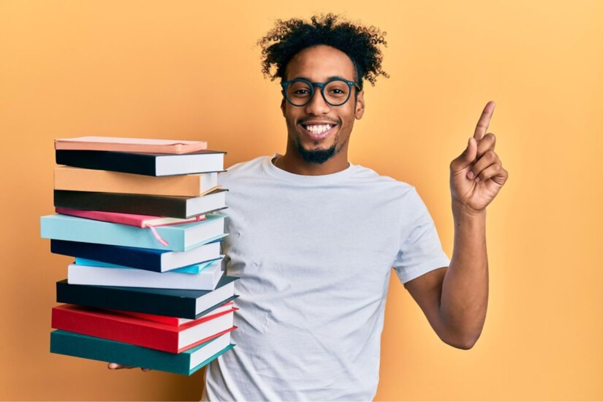 Um estudante equilibra uma pilha de livros em uma mão. Com a outra ele aponta para cima enquanto sorri