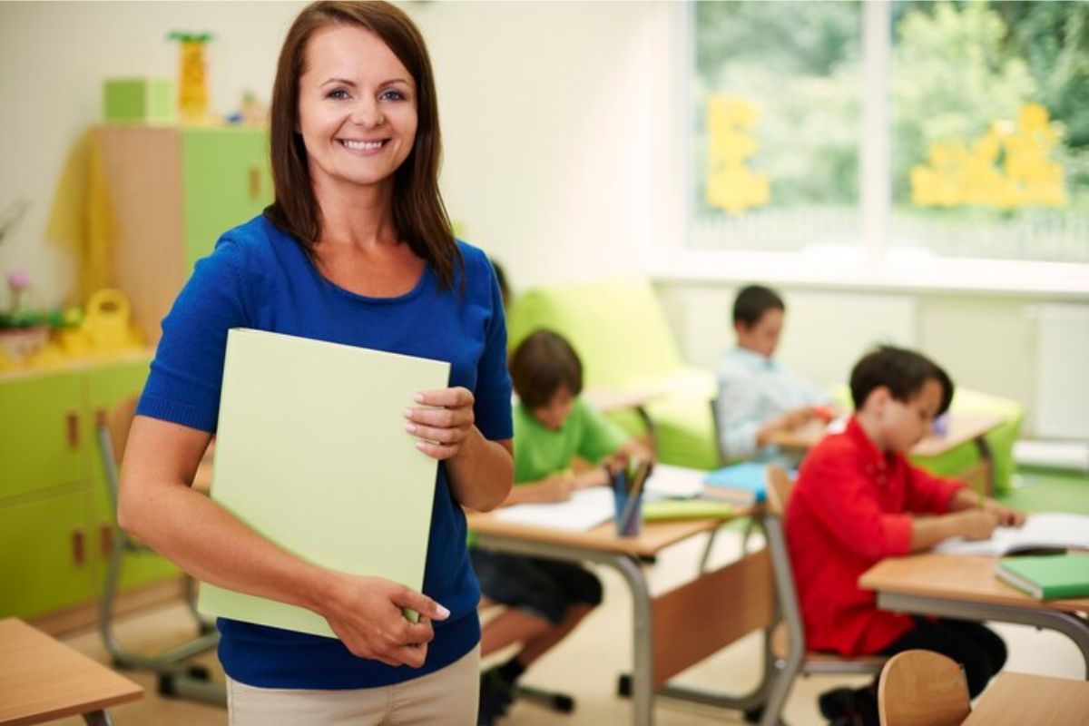 Uma professora segura uma pasta e sorri para a foto. Ao fundo é possível ver alguns alunos sentados estudando
