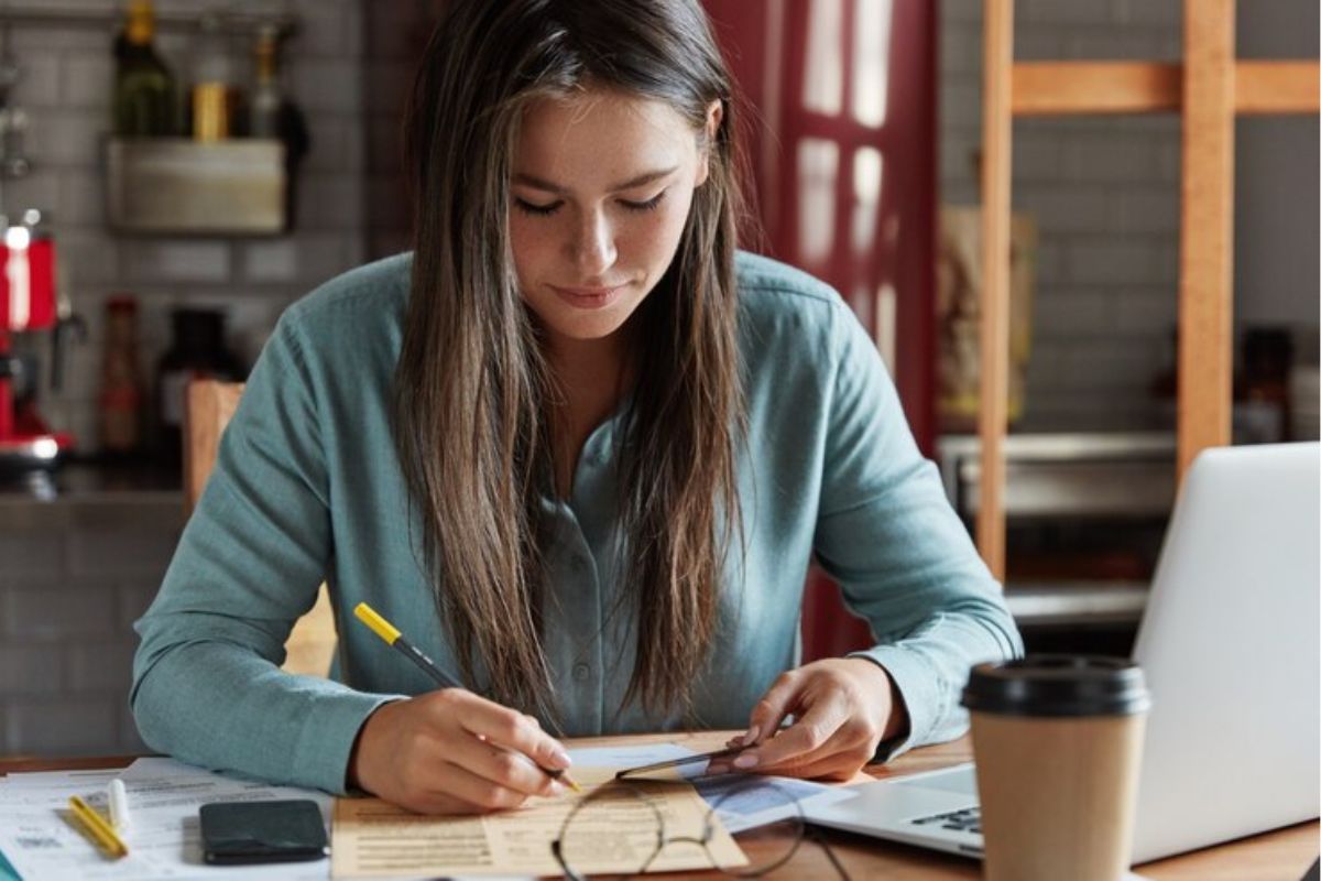Uma aluna estuda em frente ao seu notebook enquanto marca uma passagem do livro com uma caneta verde.