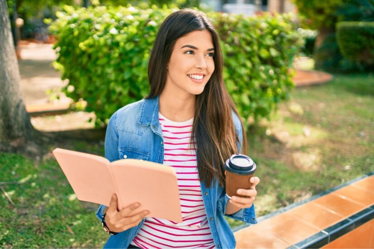 Uma aluna com um copo de café na mão e um caderno na outra sorri no jardim da faculdade
