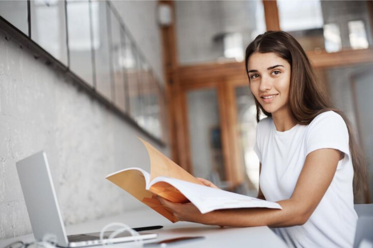Uma aluna com um livro aberto em frente ao notebook onde ela vê como funciona o FIES Segundo Semestre 2025