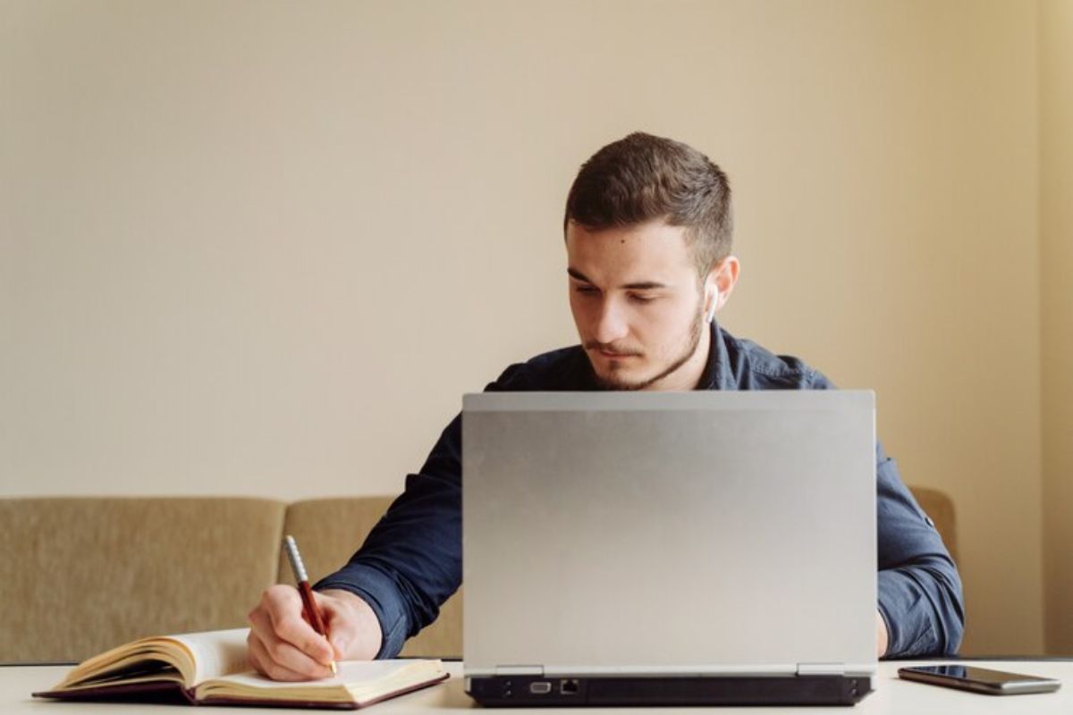 Um aluno sentado em frente ao seu notebook. Ele faz anotações em seu caderno sobre o pagamento do FIES 2025