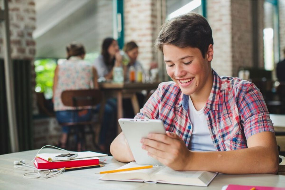 Um aluno com o tablet na mão confere o Resultado ENEM 2025, ele está sorrindo. Na mesa é possível ver um livro e alguns materiais