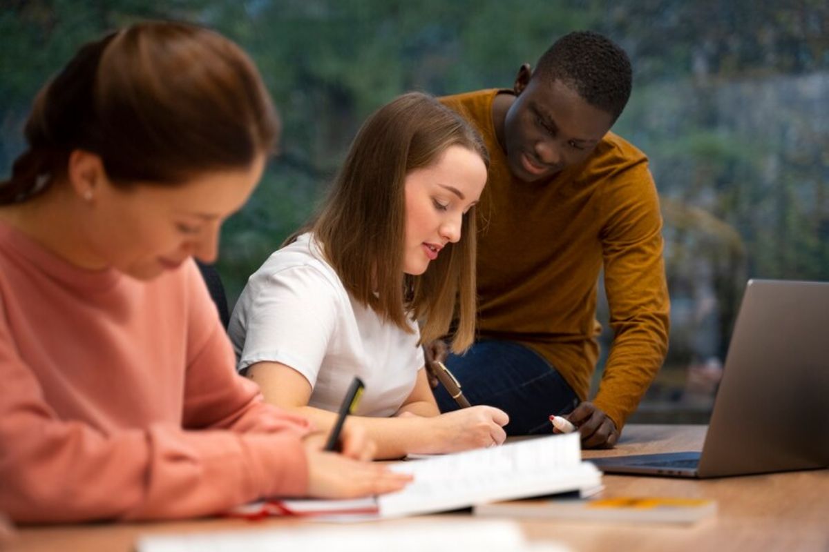 Três alunos em uma sala de aula. Um deles está sentado na mesa enquanto duas alunas escrevem no caderno