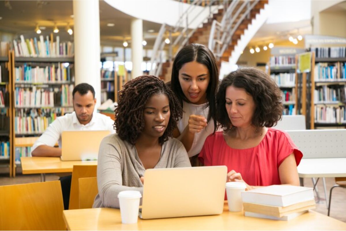 Um grupo de três mulheres dentro da biblioteca conversam enquanto olham para o notebook na mesa. Duas estão sentadas e uma fala por trás delas