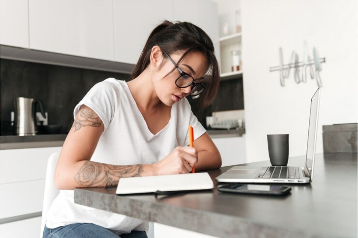 Uma mulher que tem tatuagens no braço estuda em frente ao seu notebook. Ela anota informações em seu caderno
