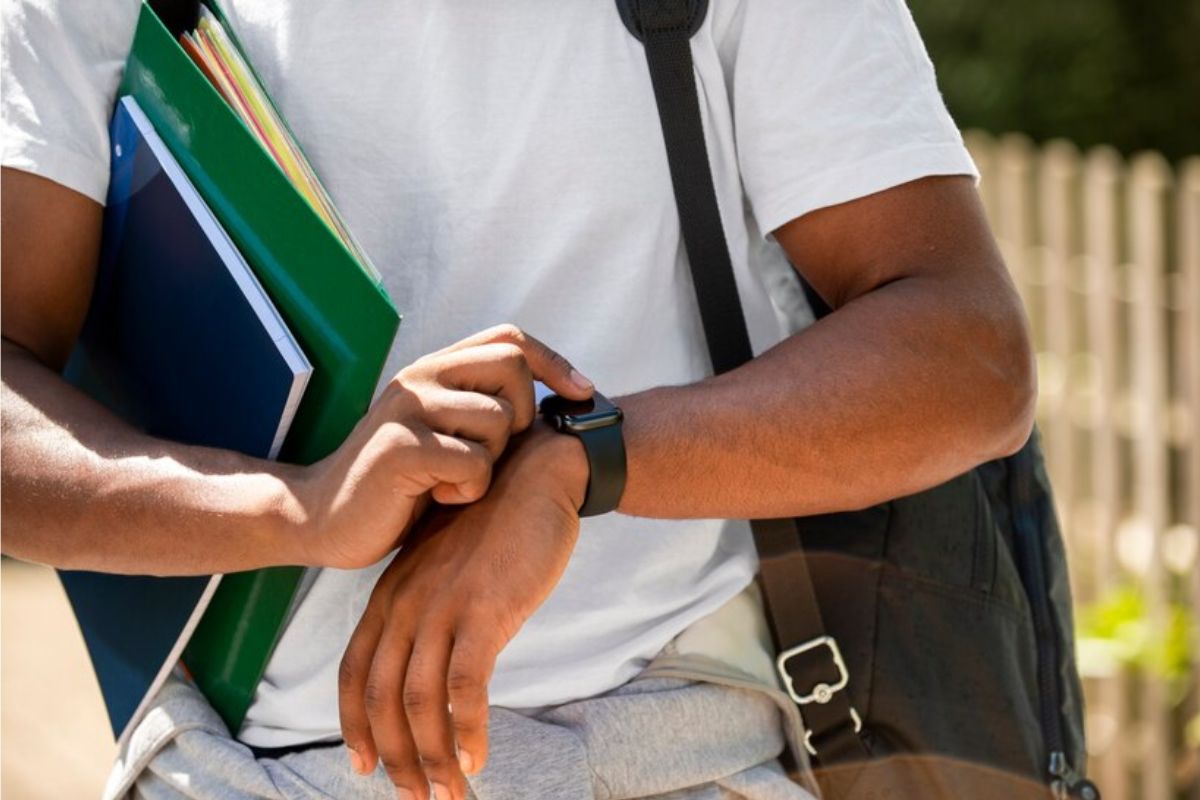 Um aluno está segurando algumas pastas embaixo do braço enquanto mexe em seu smartwatch