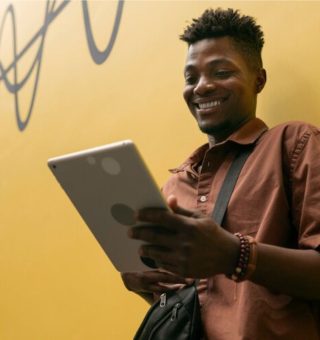 Um aluno encostado em uma parede amarela olha para o tablet que está em sua mão. Ele está vendo as Regras FIES 2025
