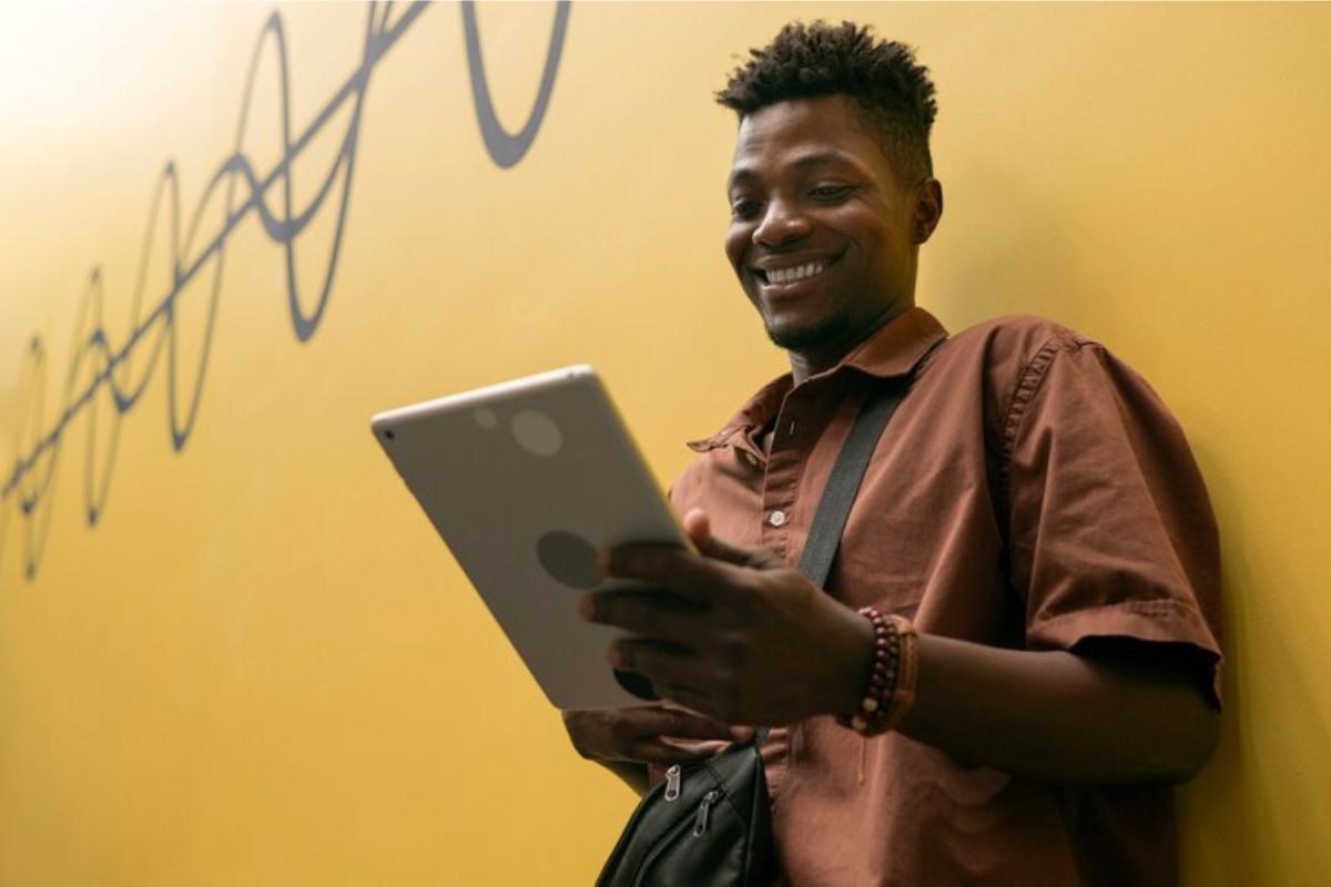 Um aluno encostado em uma parede amarela olha para o tablet que está em sua mão. Ele está vendo as Regras FIES 2025