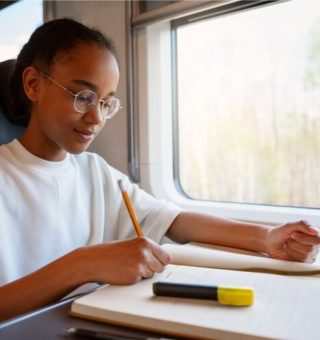 Uma menina está estudando dentro do ônibus. Apoiada em uma mesa, ela escreve em um livro com questões de Filosofia no ENEM 2025