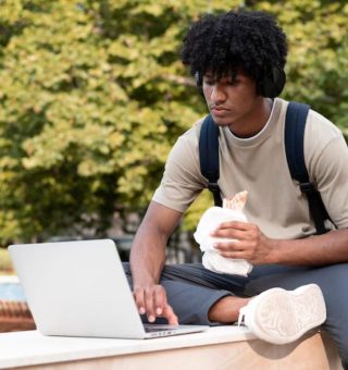 Um jovem sentado na mureta da universidade com um salgado na mão. Ele consulta em seu notebook os 7 cursos mais difíceis de passar no Prouni 2025
