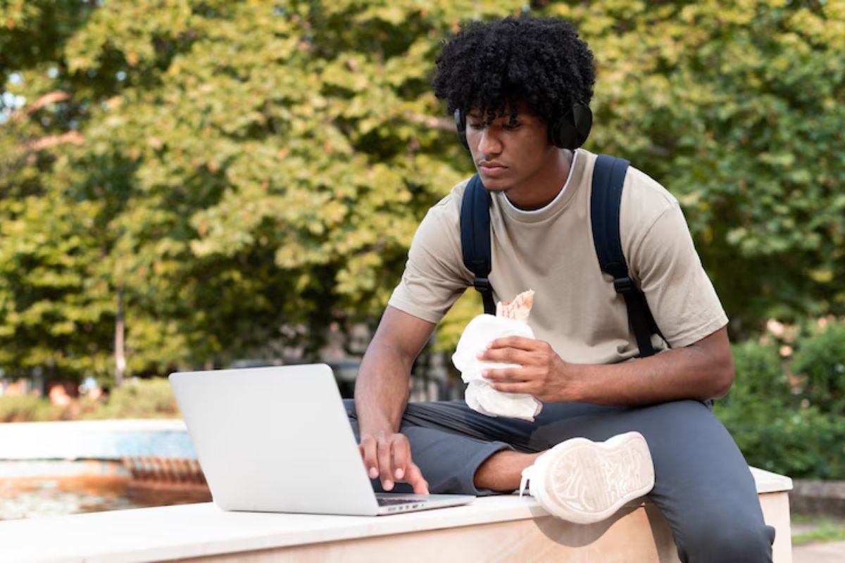 Um jovem sentado na mureta da universidade com um salgado na mão. Ele consulta em seu notebook os 7 cursos mais difíceis de passar no Prouni 2025