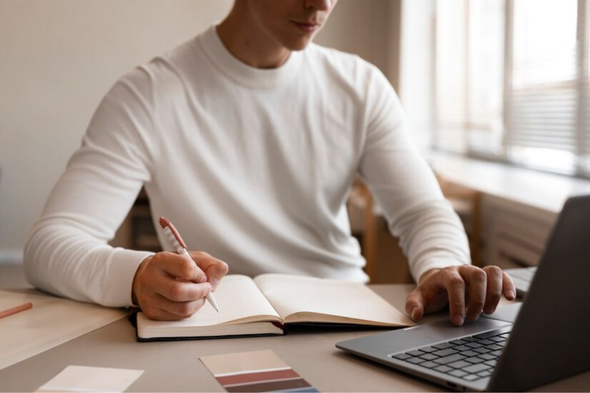 Um aluno em frente ao notebook faz anotações em seu caderno. Ele estuda para Biologia no ENEM 2025