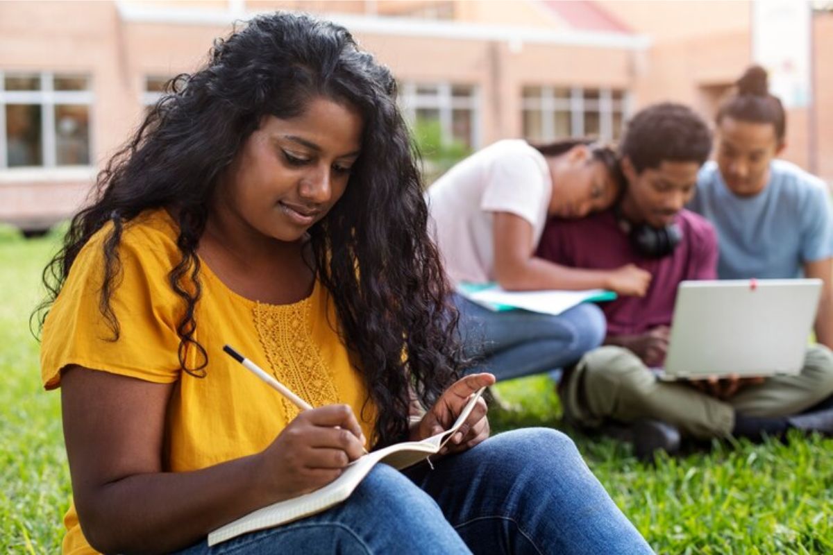 Uma aluno sentada no jardim da universidade faz anotações no caderno. Ao fundo, três amigos olham em um notebook as Regras FIES 2025