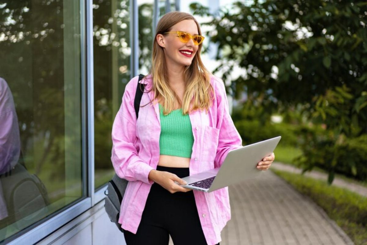 Uma mulher segura seu notebook na mão. Ela está vestindo uma grande blusa de frio rosa e caminha entre os campus da universidade enquanto confere a Lista de Espera ProUni 2025