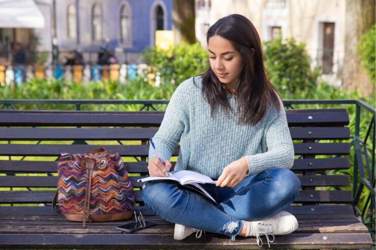 Uma aluna sentada ao ar livre enquanto escreve em seu caderno. Ao seu lado está a mochila