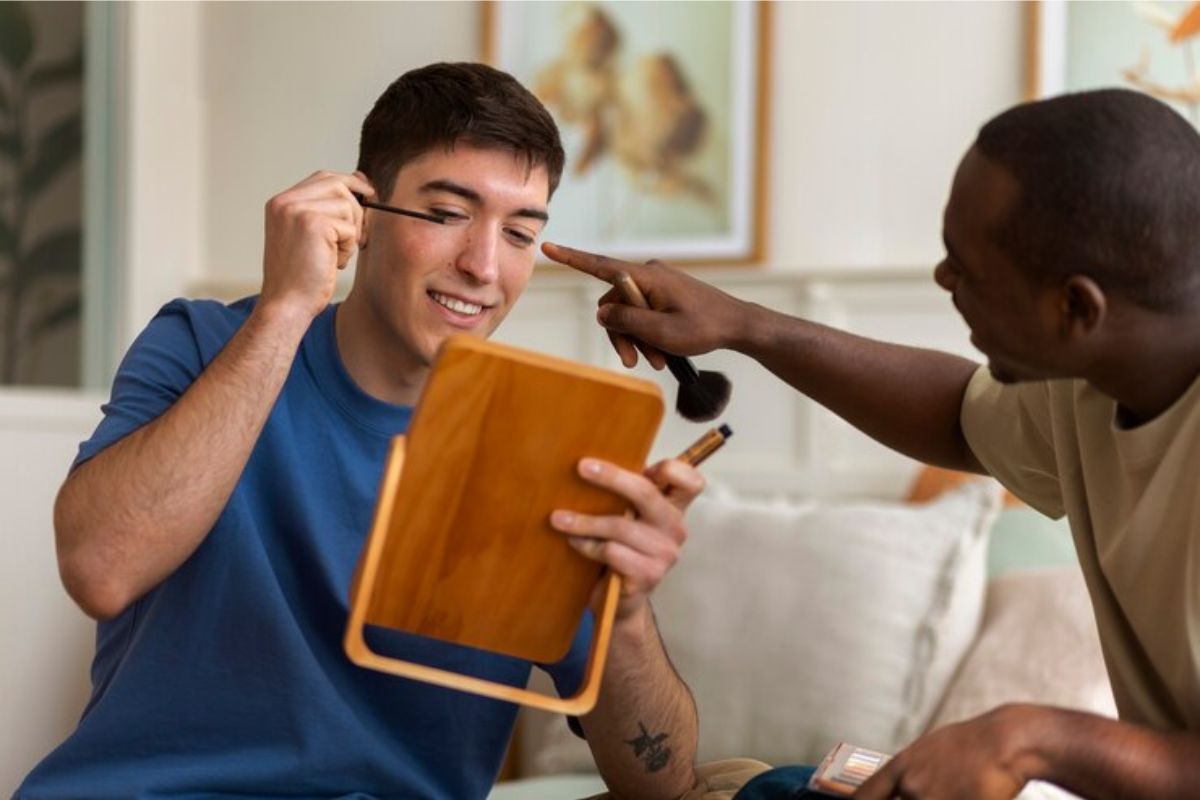 Um homem passando maquiagem olhando no espelho. Um amigo aponta para ele enquanto fala Como Usar Nome Social no ENEM 2025