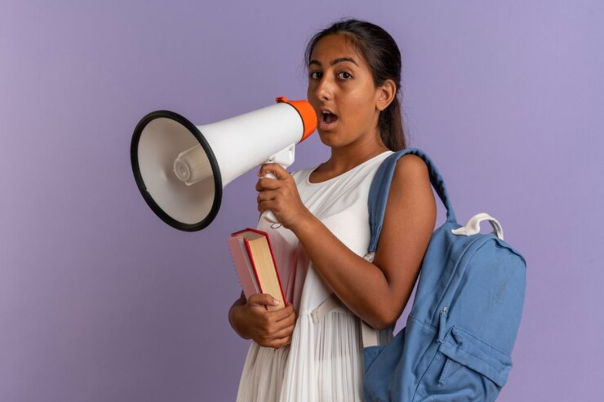 uma estudante segura um megafone que leva à boca. Com a outra mão ela segura um livro e sua mochila está pendurada em seu ombro por uma alça