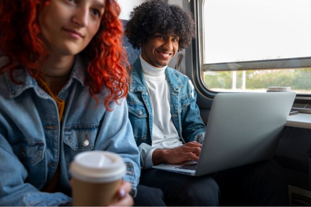 Dois estudantes no fundo do ônibus. Um menino está com o notebook no colo e a menina segura um copo de café
