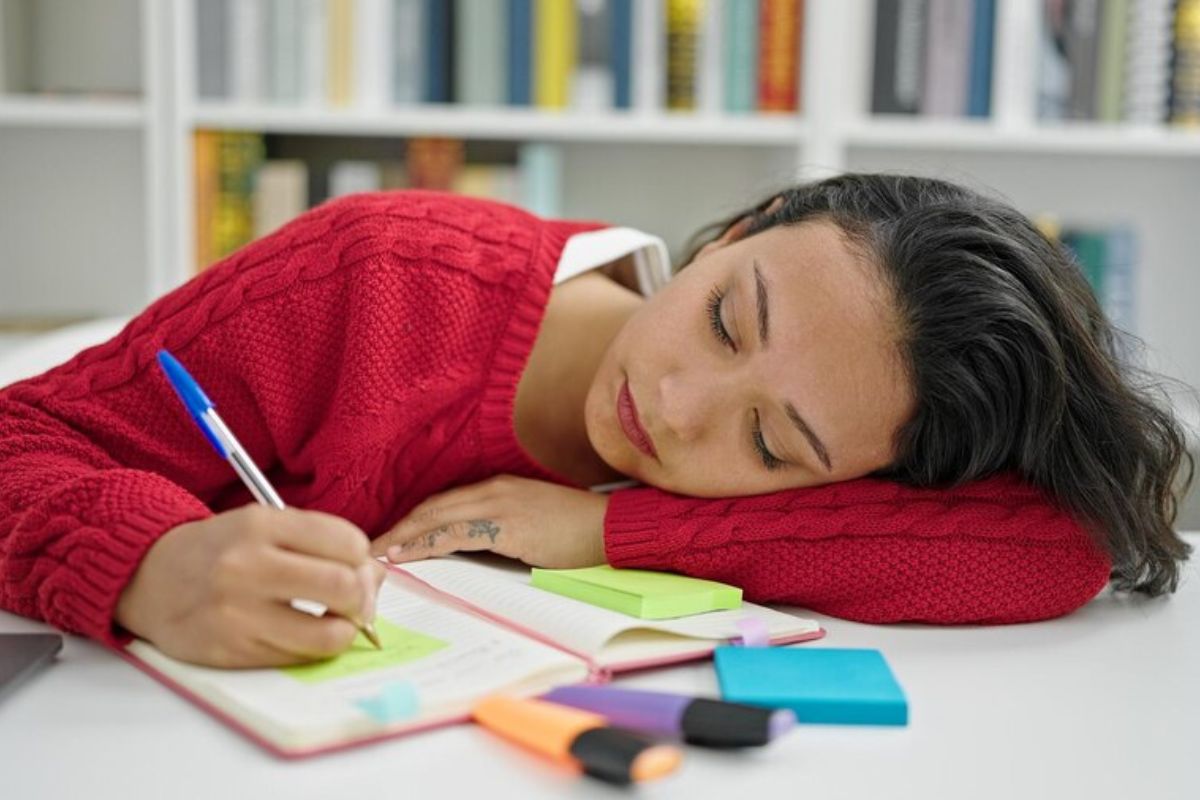 Uma menina com a cabeça deitada na mesa faz anotações em seu caderno. Ela estuda na sua casa para passar na prova da ETEC São Paulo 2025