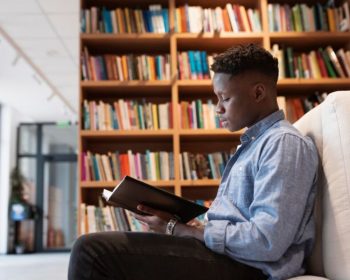 Um estudante lê na biblioteca um documento de como comprovar informações ProUni 2025. Ele está sentado e ao fundo há uma estante de livros